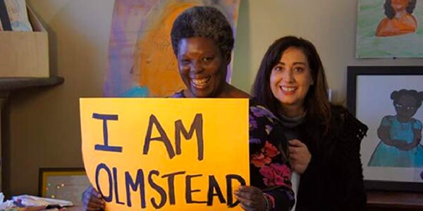 Lois Curtis smiles, holding a sign reading âI am Olmsteadâ while standing with another woman in front of a wall of paintings.