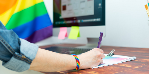 A worker wearing a rainbow bracelet writes on a piece of paper at a desk. A Pride flag is displayed next to a computer.