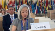 ILABs deputy undersecretary, Thea Lee, speaks at a table with Etas-Unis name card. Behind her are several international flags. 