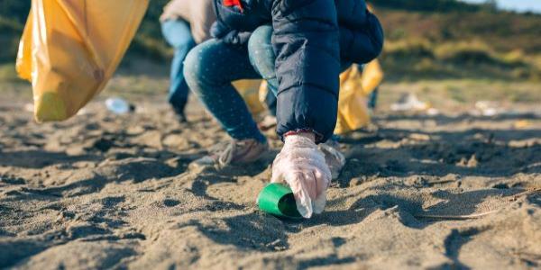 A person picks up trash on a sandy beach wearing latex gloves, a blue jacket and jeans. 