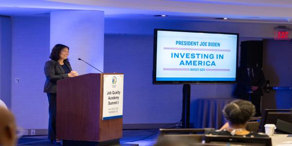 Acting Secretary Julie Su speaking behind a podium with a Job Quality Academy Summit signage and a projection that reads President Joe Biden Investing in America. 