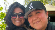 Two women smile for the camera while at a park on a sunny day. One wears an LA ballcap. One wears tinted sunglasses. 