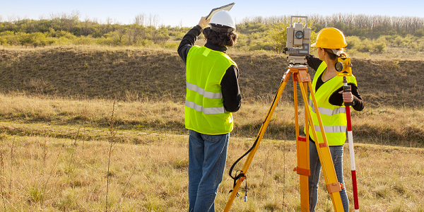 Two surveyors stand outside in a field with their equipment. 