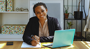 A woman sitting at a desk with a laptop, paper and pen smiles at the camera.