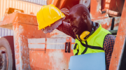 A man sits on a powered haulage vehicle on a hot day, wearing safety gear and wiping sweat from his forehead.