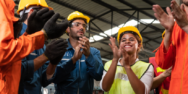 A group of workers in hard hats and safety equipment stands in a circle, smiling and clapping.