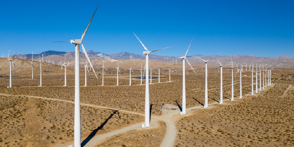 Rows of enormous wind turbines spin in an arid field beneath a blue sky.