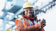 Standing in front of a crane on a worksite, a woman in protective vest, gloves, hardhat and goggles smiles at the camera. 