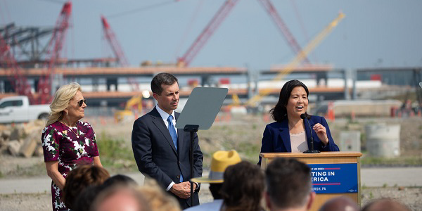 Acting Secretary Julie Su speaks at a podium in front of an airport construction project. First Lady Jill Biden and Secretary Pete Buttigieg look on. Cranes rise in the distance behind them.