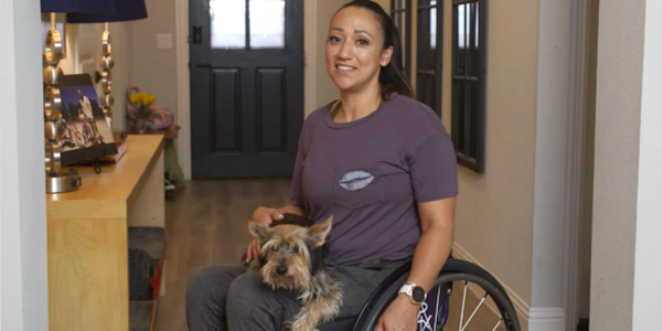 Vanessa Ross in her home. She uses a wheelchair and is holding a small dog.