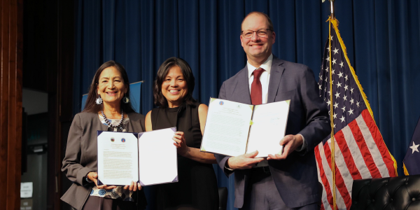 On a stage in front of an American flag, Interior Secretary Deb Haaland, Acting Secretary of Labor Julie Su and EPA Chief of Staff Dan Utech hold up a signed memorandum. 