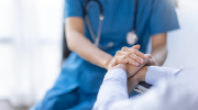 Close-up shot of a person in scrubs with a stethoscope holding the hands of a seated adult.