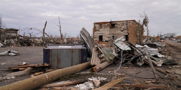 Tornado damage. Stripped trees rise toward grey winter skies. A downed telephone pole, crumpled sheet metal & other rubble are scattered across a lot in front of a battered house with blasted windows & siding. Photo by Dominick Del Vecchio, FEMA
