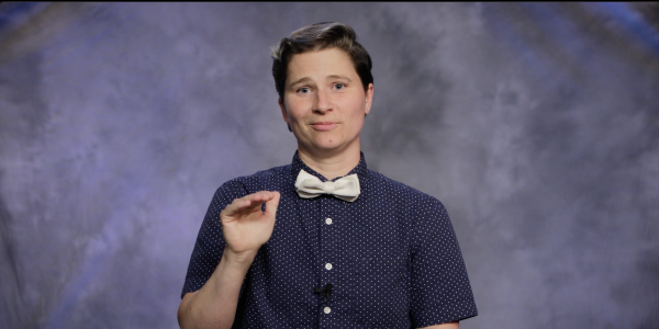 A person with short hair and a bowtie stands in front of a grey backdrop, facing the camera, with one hand raised to emphasize a point. 