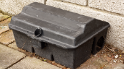 A boxy plastic rat trap sits on the sidewalk near a building's exterior. 