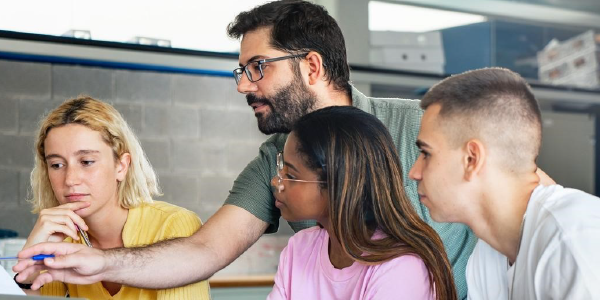 Three young people look at a computer while an adult gestures at the screen to illustrate a point.