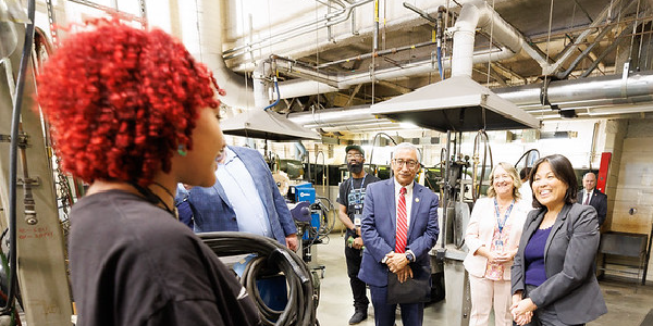 Secretary of Labor Julie Su, Rep. Bobby Scott and others chat with a young professional working in a factory setting. Ducts are visible overhead. 