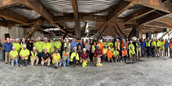 A large group of people â many wearing hard hats and safety vests â pose for a photo in a building under construction.