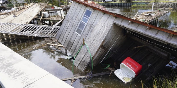 A shed lists at an angle, having been blown into a canal. Debris surrounds it, including split wood, an unconnected railing and an upended cooler. Source: Robert Kaufmann, FEMA