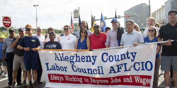 Ahead of a parade, Acting Secretary Su stands with a dozen people holding a banner reading 'Allegheny County Labor Council AFL-CIO, Always working to improve jobs and wages.'