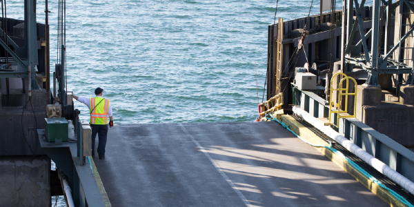 A longshore dock worker in a reflective vest stands at the edge of a ferry boat pier, gazing out over the water.