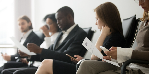 Five professionally dressed workers of different ethnicities sit in a waiting room, holding resumes and cellphones.