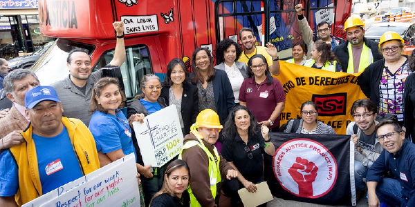 Julie Su poses for a photo with workers and advocates who hold signs with messages supporting worker rights. The workers, some in uniforms and safety gear, smile, and some raise their fists in a symbol of solidarity. A bus behind the group is painted with