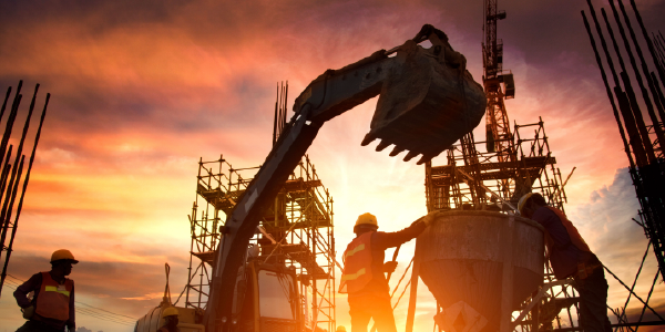 A construction site appears in silhouette before the setting sun. Workers, scaffolding and diggers are visible.