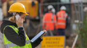 A woman construction worker in safety gear talks on a phone while consulting a tablet. Coworkers in orange vests and hardhats are visible in the background. 