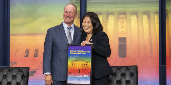 Acting Secretary Su and Gerald Bostock smile in front of a backdrop featuring the Supreme Court in the colors of the diversity pride flag, holding a signed poster commemorating the Hall of Honor induction. 