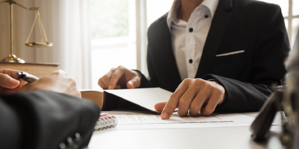 Two men in business suits discuss a document placed between them. The scales of justice are visible in the background. 