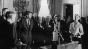 Black-and-white photo shows Esther Peterson, John F. Kennedy and others gathered in the White House.