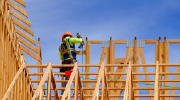 Viewed from below, a man in safety gear stands on a house under construction, using a nail gun to assemble the frame.