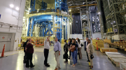 Men and women in professional clothing stand in a warehouse setting where large aerospace equipment is visible in the background.