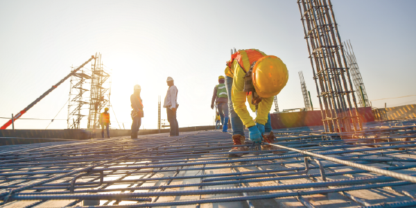 Construction workers in safety vests and hard hats bend over scaffolding atop a rising construction site.