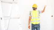 A worker wearing a high visibility vest and hardhat, seen from behind, touches up a white wall with a paint brush.