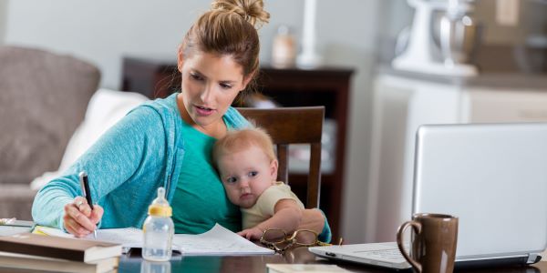 A young woman holds a baby in one hand while writing in a notebook, with a laptop open on the table in front of her. 