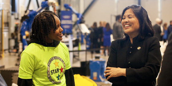 In a warehouse, Acting Secretary Su chats with a woman wearing a bright tee shirt. Both are smiling. 