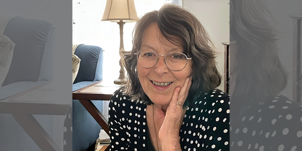 A woman in glasses and a polka-dot shirt smiles at the camera, seated in a living room.