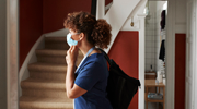 A woman in medical scrubs and a surgical mask walks through the foyer of a home.