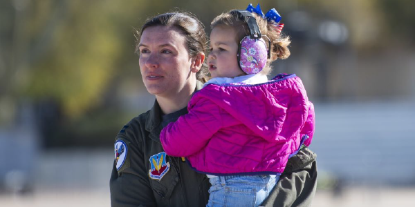 A woman in military uniform holds a toddler wearing a jacket and noise-cancelling headphones