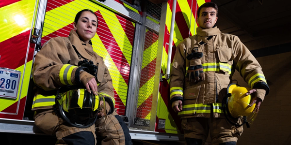 A man and woman in firefighting gear stand in front of a firetruck. U.S. Air Force Photo by Airman 1st Class Trevor Bell.