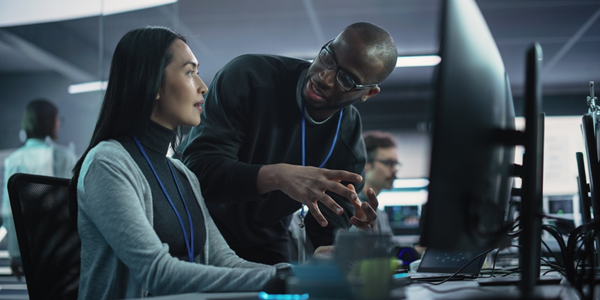 A female worker sits at a computer station, engaged in conversation with a man standing next to her. 