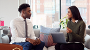 A man and woman chat in an office. The woman has a laptop open and the man listens attentively.