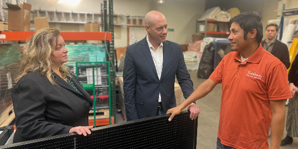 Lisa Gomez and Greg Landsman examine a solar panel in a workshop while chatting with a man in an orange shirt with a Sustainergy logo.