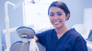 A smiling dental hygienist leans on a patient chair.