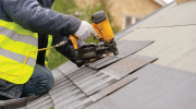 A person in safety gear attaches roof shingles to a sloped roof.