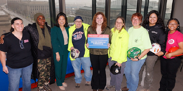 Acting Secretary of Labor Julie Su with eight women workers holding hard hats and a sign that says President Joe Biden, Investing in America.