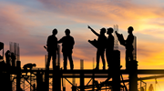 The silhouettes of several construction workers standing on scaffolding as the sun rises behind them.