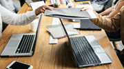 Several people work together at a large table with laptops, notebooks and files.